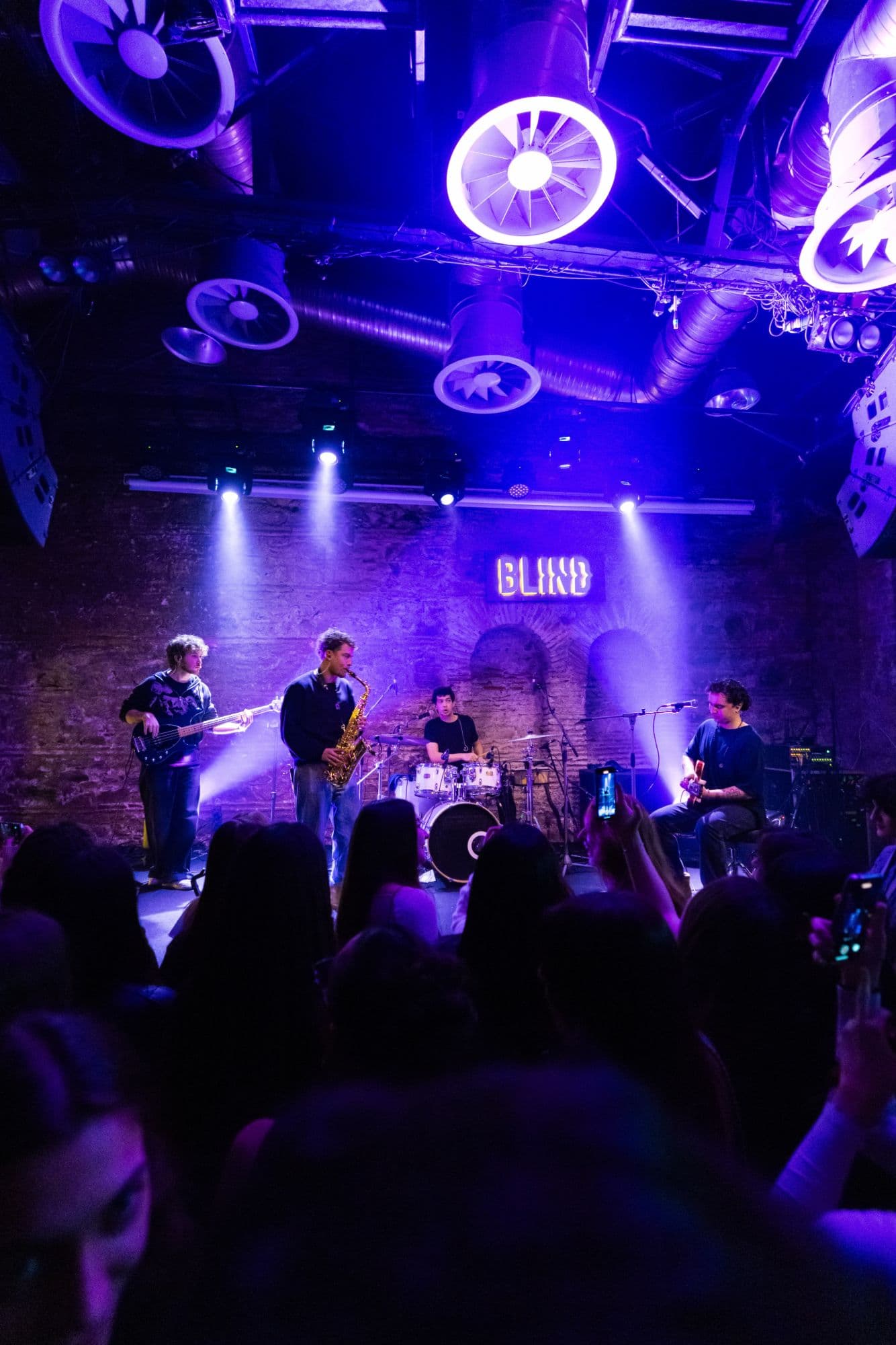 Maymun Collective playing on the stage at Blind under purple light, phones raised in the crowd.